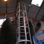 Black chemical storage tank with ladder access and blue scissor lift