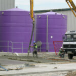 Two purple tanks outside next to each other while two workers dig a hole nearby with a crane